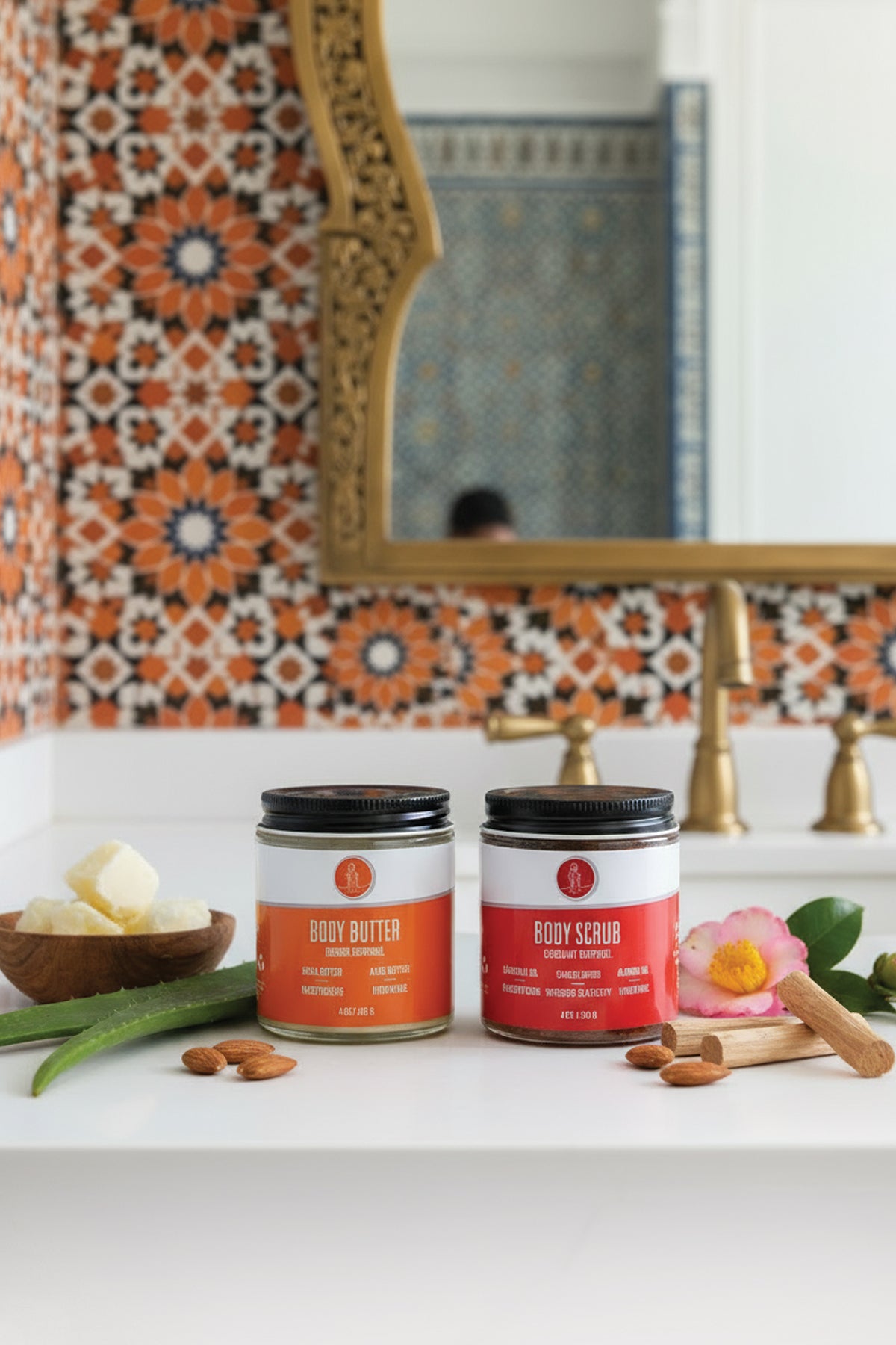Two jars of body butter and scrub on a bathroom counter with decorative tiles and mirror in the background.