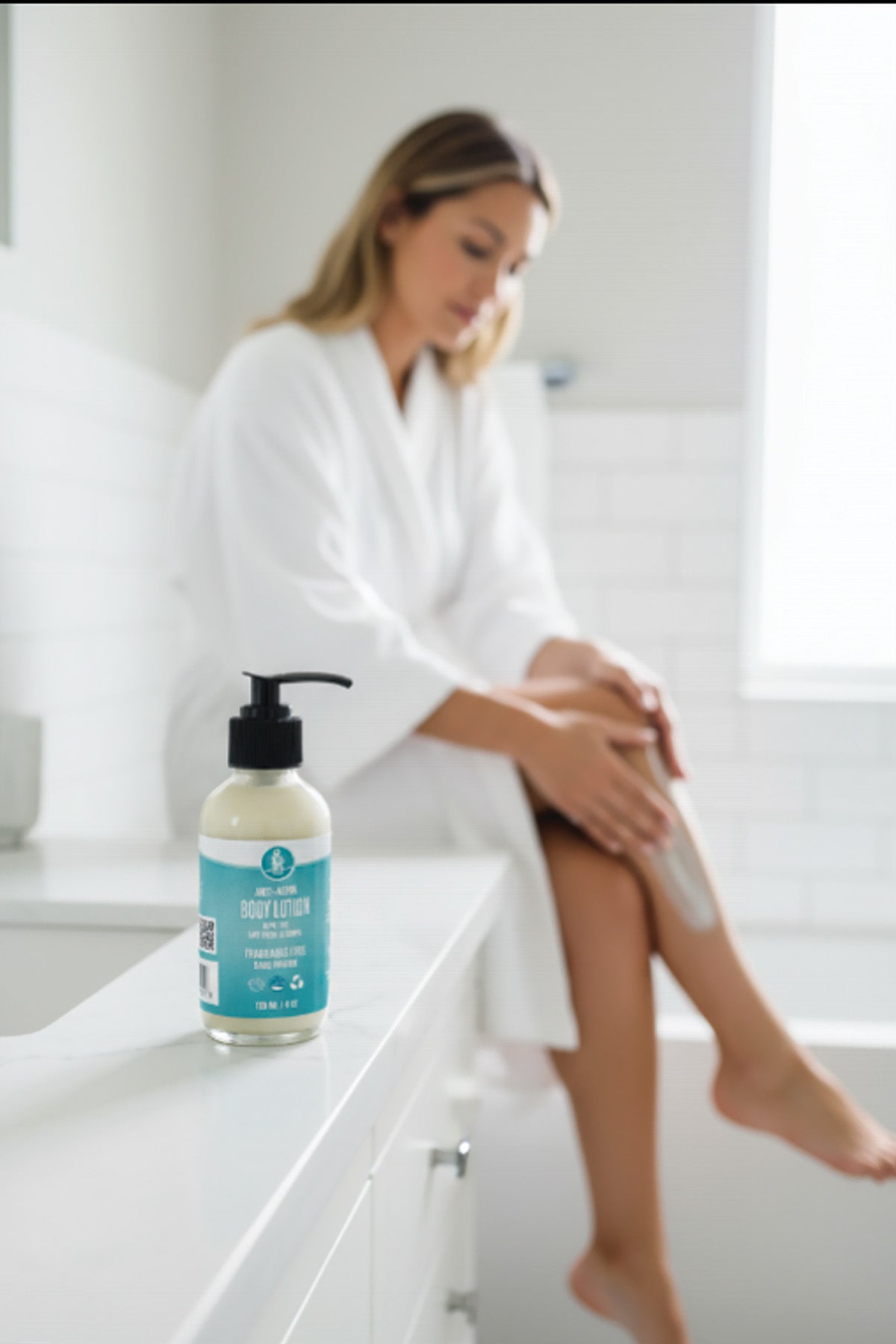 Woman in a bathrobe sitting on a bathtub with a bottle of body lotion next to her.