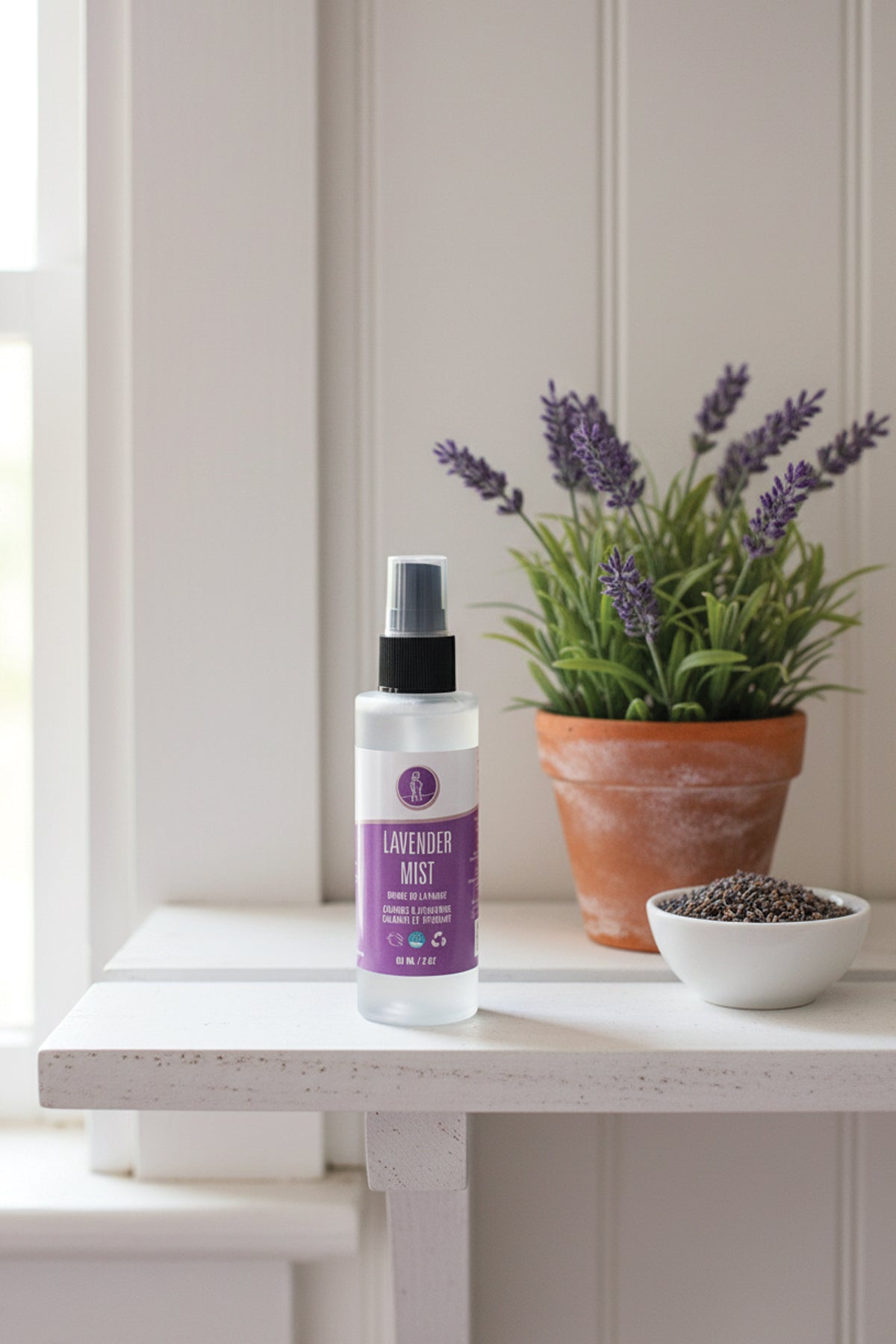 Lavender mist spray bottle with a plant and small bowl on a white surface.