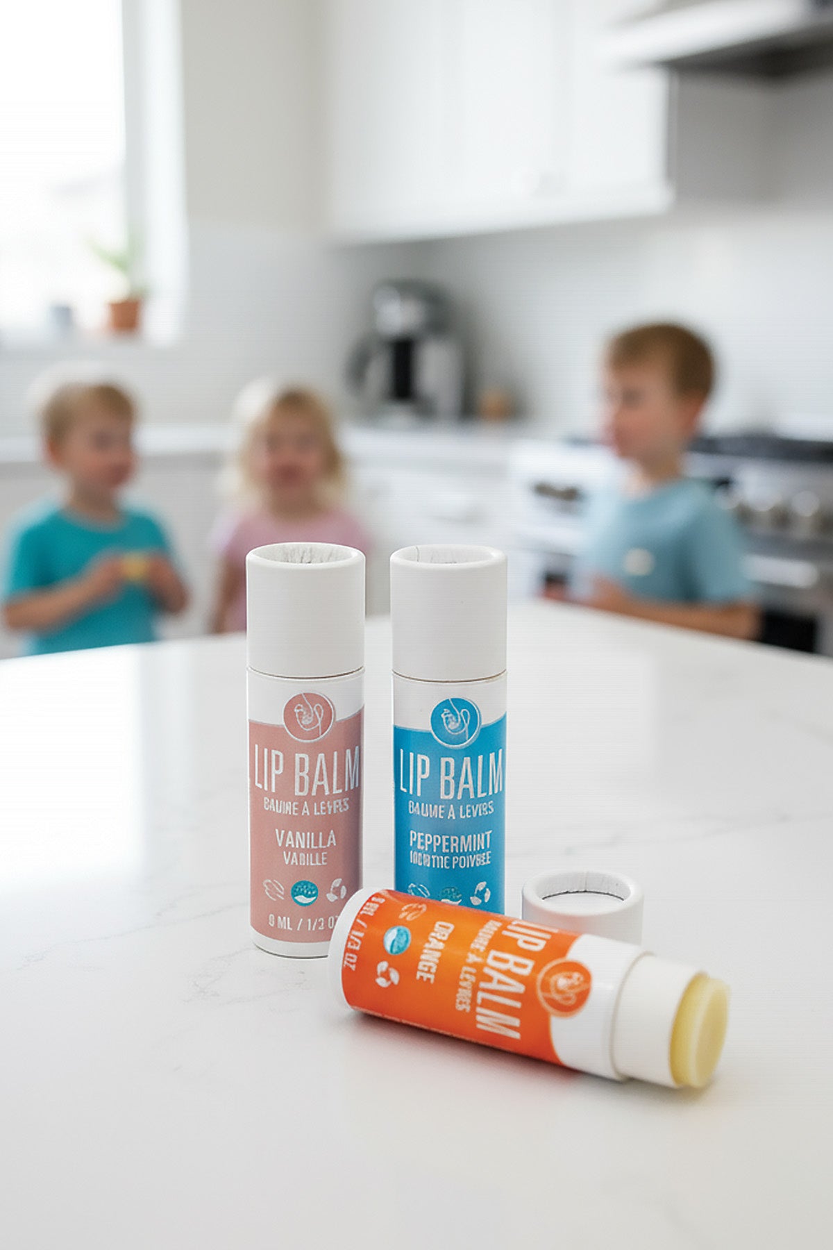 Three lip balm containers on a kitchen counter with children in the background