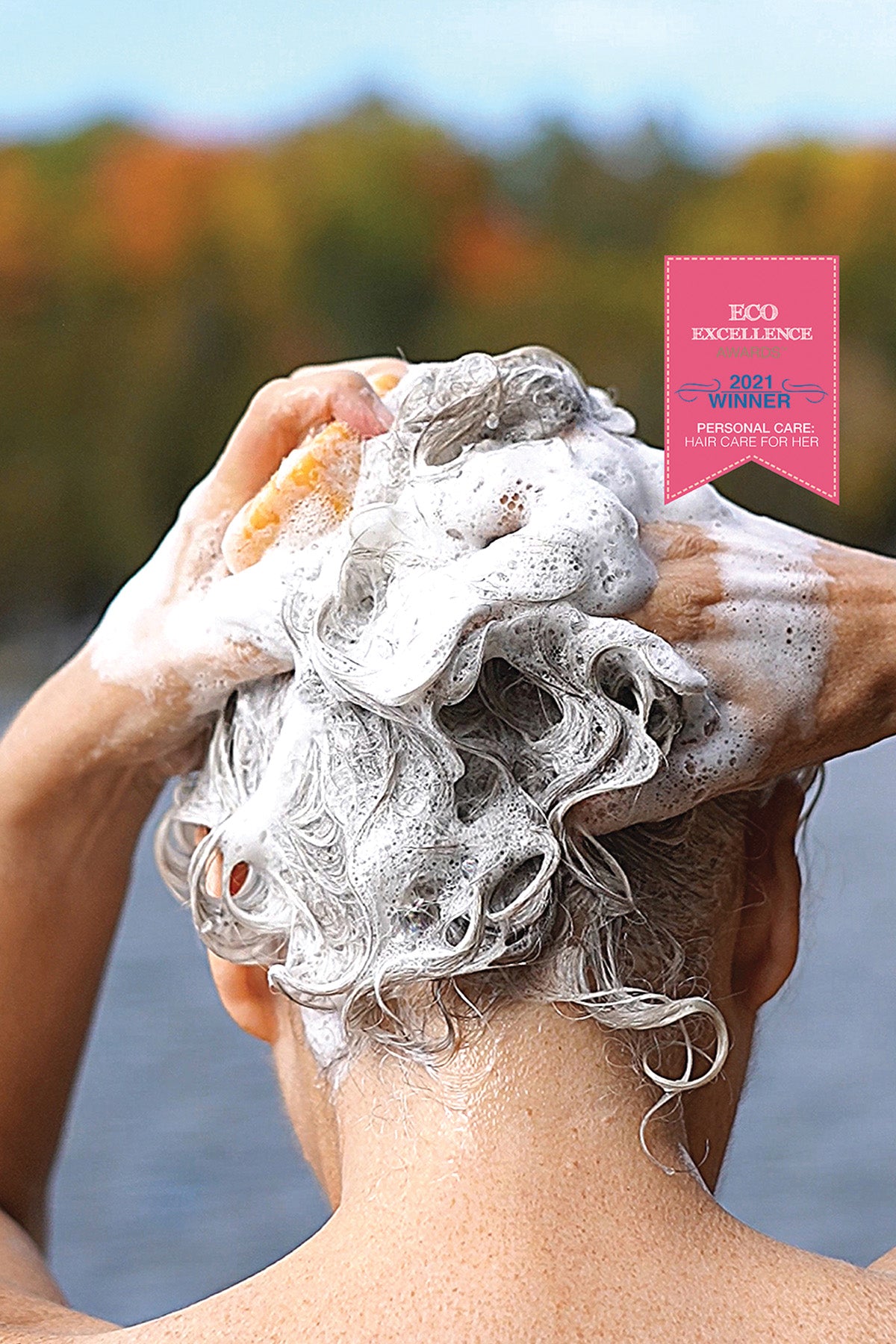 Person washing hair with soap suds outdoors, with a product label in the corner.
