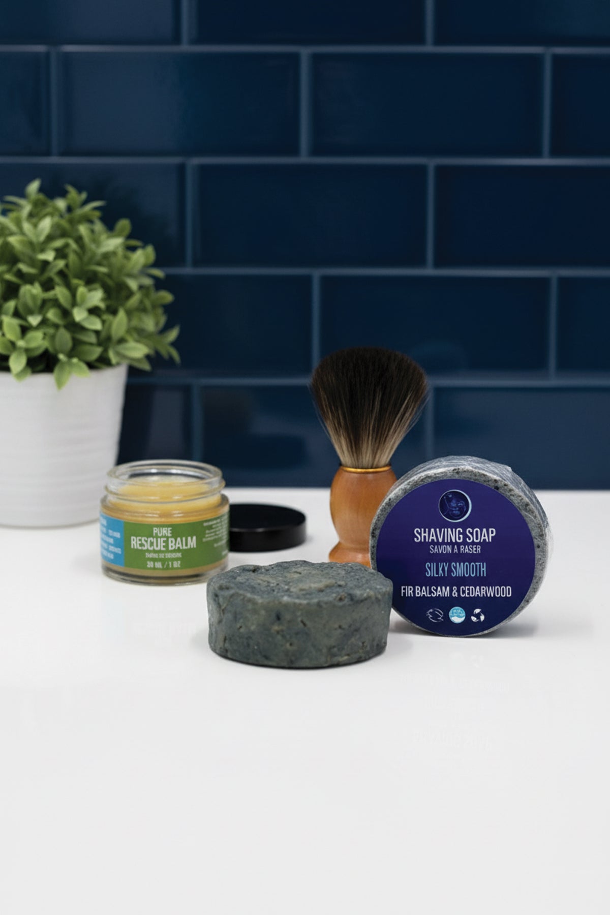 Bathroom setting with shaving products including a jar, soap, and brush against a blue tiled wall.