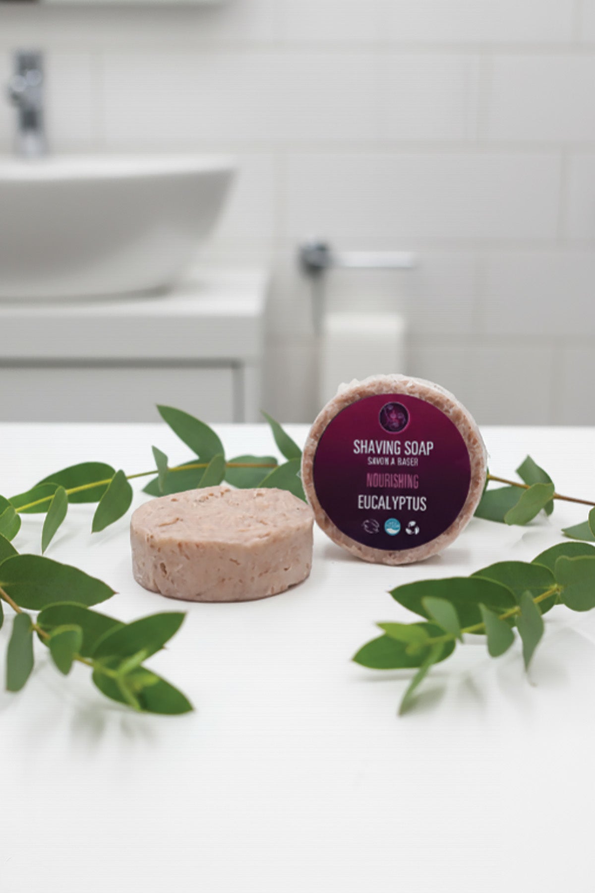 Two shaving soaps with eucalyptus leaves on a bathroom counter.